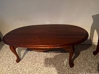 Front view of oval wood coffee table with curved legs and cherry finish on carpet.