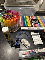 Photo showing clear plastic container, colored markers in circular holder, Bright Ideas colored pencils tin case, glue stick, protractor, clip binder, sharpener, erasers, pencils, ruler and Sharp EL-244W calculator