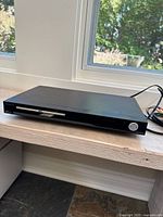 Front and top view of black Sylvania DVD/Blu-ray player on wooden surface with natural light from windows.