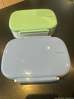 Two rectangular plastic food storage containers with white bases, periwinkle and light green locking lids, placed side by side.
