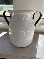 Front view of white ceramic pot with black metal handles showing rounded body and wide top rim.