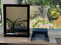 Rectangular wall mirror with dark brown wood frame and small blue stool positioned in front, shown on wood table with outdoor background.