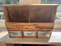 Front view of wooden wall-mounted cabinet showing two open compartments at the top separated by a central divider and three metal-front drawers with metal handles below.