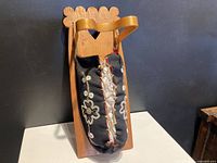 Front view of cradle board showing scalloped wooden top, leather strap, and black fabric pouch with floral beadwork.