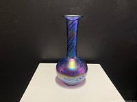 Full view of Arte Varges iridescent glass vase with bulbous base and long neck, blue-purple colors with swirling pattern, on white pedestal against dark background.