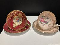 Two fine bone china tea cups with matching saucers, one red with gold accents and one pale pink with floral pattern and gold accents.