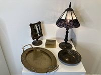 Photo of overall lot arrangement showing brass tray, slag glass lamp, brass mirror, brass bowl, and brass trinket box on a white table.
