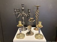 Five brass candle holders shown together, including candelabra, two porcelain-style stem holders, twisted spiral stem holder, and flower-shaped holder.