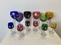 Full view showing six colored cut to clear crystal wine glasses and six matching cordial glasses on white surface.