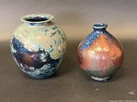 Two signed Raku pottery vases side-by-side showing their size difference and glaze color variations under soft lighting.