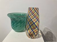 Two signed art glass vases side by side on a white surface, showing one green swirled vase with flared rim and one colorful criss-cross patterned vase.