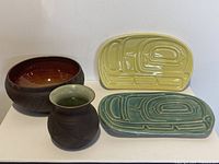 Four pottery pieces: two brown pottery with tribal and geometric designs, one signed on the bottom, and two ceramic plates with raised tribal designs in yellow and green.