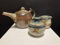 Front and side view of signed studio pottery tea pot, creamer, and sugar bowl on white surface