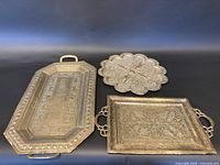 Group photo showing three brass trays with detailed embossed designs, illustrating size and style differences.