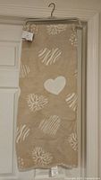 Full view of the beige throw blanket with white heart and abstract designs, hanging on a hanger.