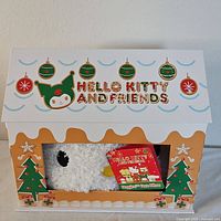 Photo of a closed holiday-themed box shaped like a gingerbread house with Hello Kitty and Friends branding containing the face pillow.