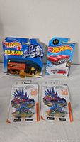 Image of four Hot Wheels die cast cars in packaging: Haulers truck with orange tank, red and white car #75, and two identical Hot Wheels id dinosaur design cars.