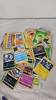 Pile of assorted Pokémon trading cards showing various characters and card designs spread on white surface.