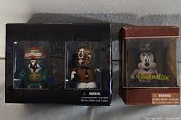 Photo of three Disney Vinylmation figures in original boxes: one single Mickey Mouse figure in a brown box and a boxed set containing two figures, one green with Disney transportation theme, another brown with animal features.