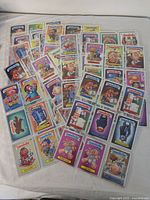 Overview of 16 Garbage Pail Kids trading cards spread out, each in protective plastic sleeves. Cards display colorful cartoonish grotesque characters typical to the series.
