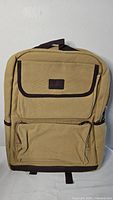 Front view of tan and brown backpack showing front zippered pocket and flap pocket with brown trim and small patch