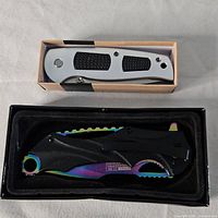 Two folding pocket knives in their boxes, showing silver handle one and black/iridescent handle one.