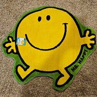 Full top-down photo of the Mr Happy shaped rug on carpet floor, showing yellow character with arms and legs outstretched and 'Mr Happy' text.