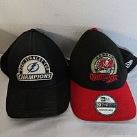 Front view of both hats showing Tampa Bay Lightning Stanley Cup Champions patch and Buccaneers logo patch with red brim.