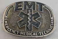 Front view of the vintage EMT belt buckle showing the Star of Life emblem, EMT lettering, and 'Emergency Medical Technician' text.