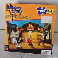Front of the puzzle box showing the Looney Tunes characters Bugs Bunny, Yosemite Sam, and Daffy Duck in a Wild West saloon scene named 'Lucky Charmin Saloon.' The box indicates 100 puzzle pieces and an age recommendation of 5-8 years.
