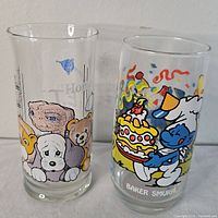 Two vintage glasses side by side, Smurf character holding a birthday cake on one and E.T. with animals on the other.