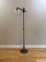 Full view of the antique metal floor lamp standing on wood floor against a light wall, showing the lamp's height, adjustable arm, and light bulb attached.