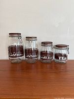 Four vintage glass canisters with metal clasp lids, labeled FLOUR, SUGAR, COFFEE, and TEA, arranged on a wooden surface against a white wall. Heights visible from left to right.