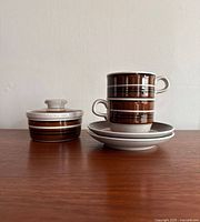 Two stacked Rorstrand Isolde coffee cups with their saucers underneath, next to a lidded sugar bowl on a wooden surface against a white wall.