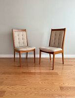 Two mid-century teak dining chairs positioned on a wooden floor against a plain wall, showing front and angled views.