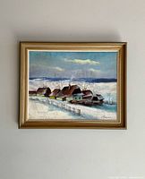 Framed oil painting showing a winter village scene with snow, houses, and fence, signed by the artist in the lower right corner.