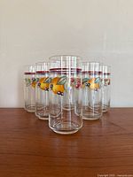 Front view of six tall drinking glasses with colorful fruit decoration on a wooden surface against a plain wall.