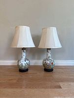 Full view of two vintage ceramic lamps with white fabric shades, standing on wooden floor near a light wall. Lamps are identical with round bases decorated with colorful bird and flower artwork.