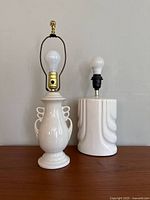 Two vintage ceramic table lamps, white finish, without shades or bulbs, shown side by side on surface against beige wall