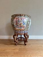 Front view of the ceramic planter showing a scene with figures under a canopy, floral and fish motifs, on a wooden stand.