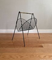 Front angled view of the vintage metal wire magazine rack, showing black wire cage and brass handle.