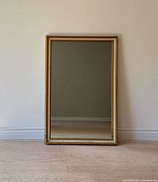 Full front view of the mirror leaning against wall on light wood floor, showing rectangular shape and gold frame.