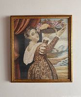 Frontal image of framed vintage needlepoint showing a woman holding a basket of flowers with cloud and curtain background.