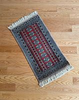 Vintage small rectangular rug from angled view showing red central field, geometric pattern, and fringed edges.