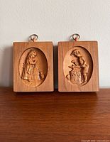 Two wood carvings on rectangular plaques, side by side, each about 5 x 4 inches with oval recessed reliefs featuring a woman churning butter and a blacksmith.