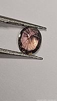 Close-up photo of the oval cut sapphire held by tweezers showing purple and pink hues.