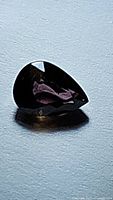 Pear-shaped faceted sapphire gemstone showing dark purple hue in ambient light, size approximately 6.16x4.43mm.