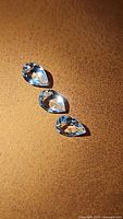 Three natural pear cut aquamarine gemstones arranged vertically on a brown surface, showing their faceted cuts and light blue color.