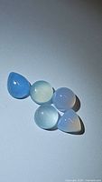 Five natural chalcedony cabochons arranged in mixed shapes and light blue to lavender colors on white background with lighting highlighting translucency and surface gloss.