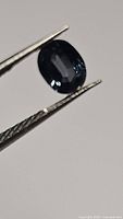 Close-up photo of an oval cut gemstone held by tweezers. The stone is a deep blue to purple color and transparent with some reflection.